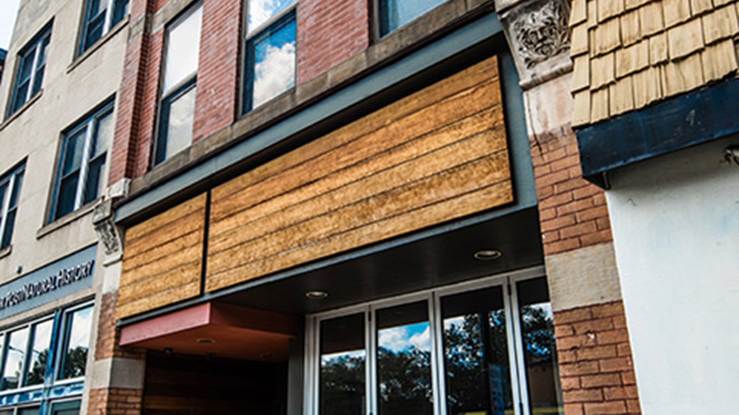 4915 Penn Ave exterior. A building facade featuring a large, blank wooden panel covering a storefront. The brick structure shows windows above, with a decorative element on the right. Reflections of clouds are visible in the windows.