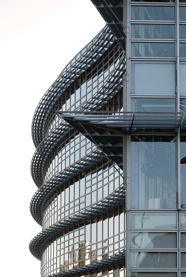 A close-up view of a modern building's exterior featuring curved glass panels and a layered design. The structure displays a series of horizontal lines and reflective surfaces, with some architectural elements extending outward.