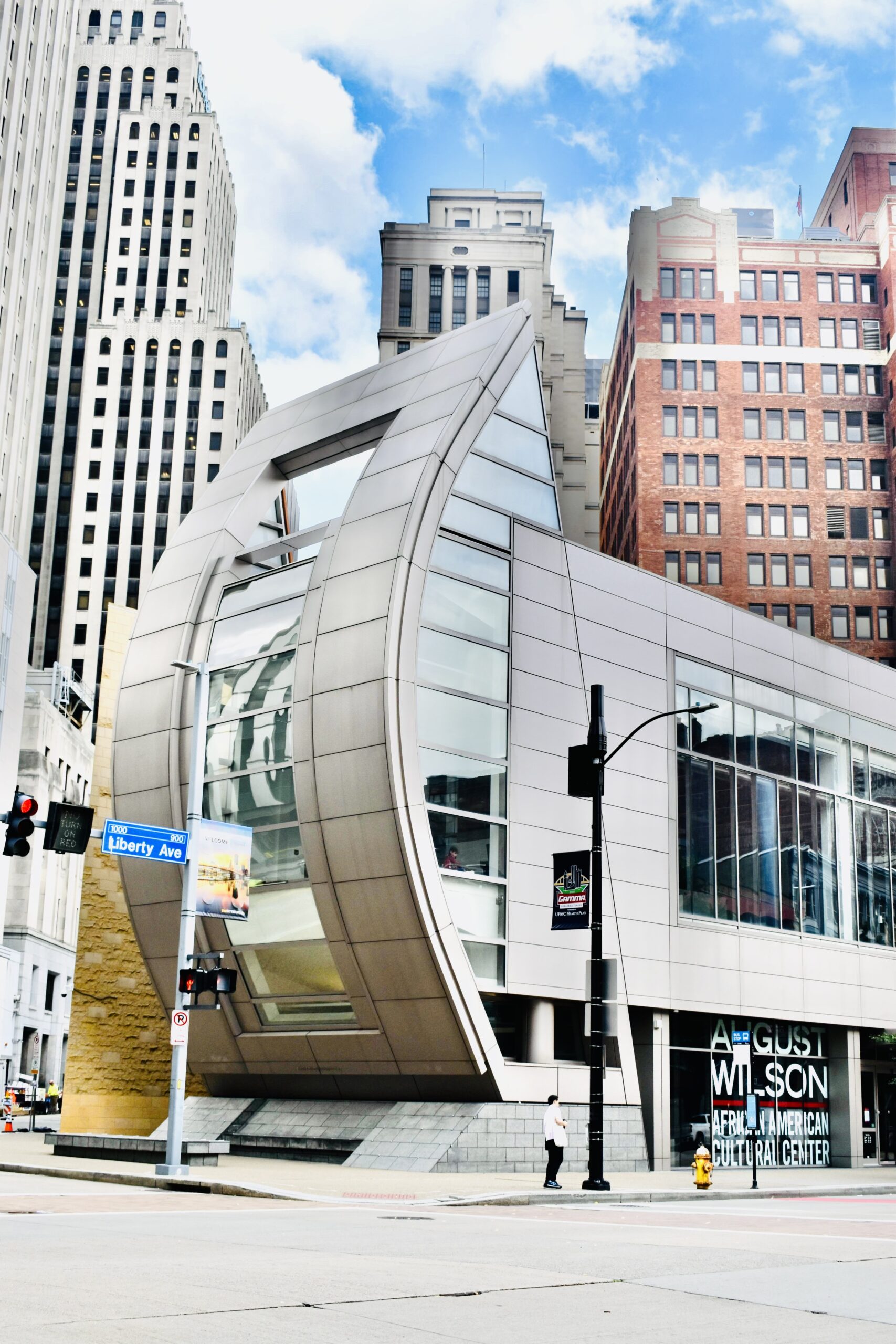 A modern building with a curved metallic façade located at a city intersection. Tall buildings surround it, and a pedestrian stands near the entrance. The sky is partly cloudy.