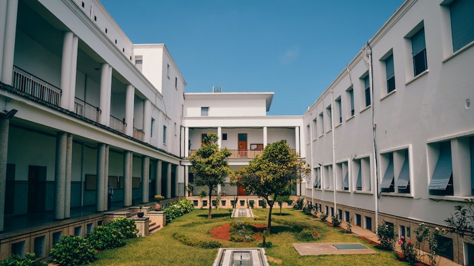A view of a courtyard surrounded by white buildings, with green grass and trees in the center. The architecture features columns and balcony spaces on the left and right sides.