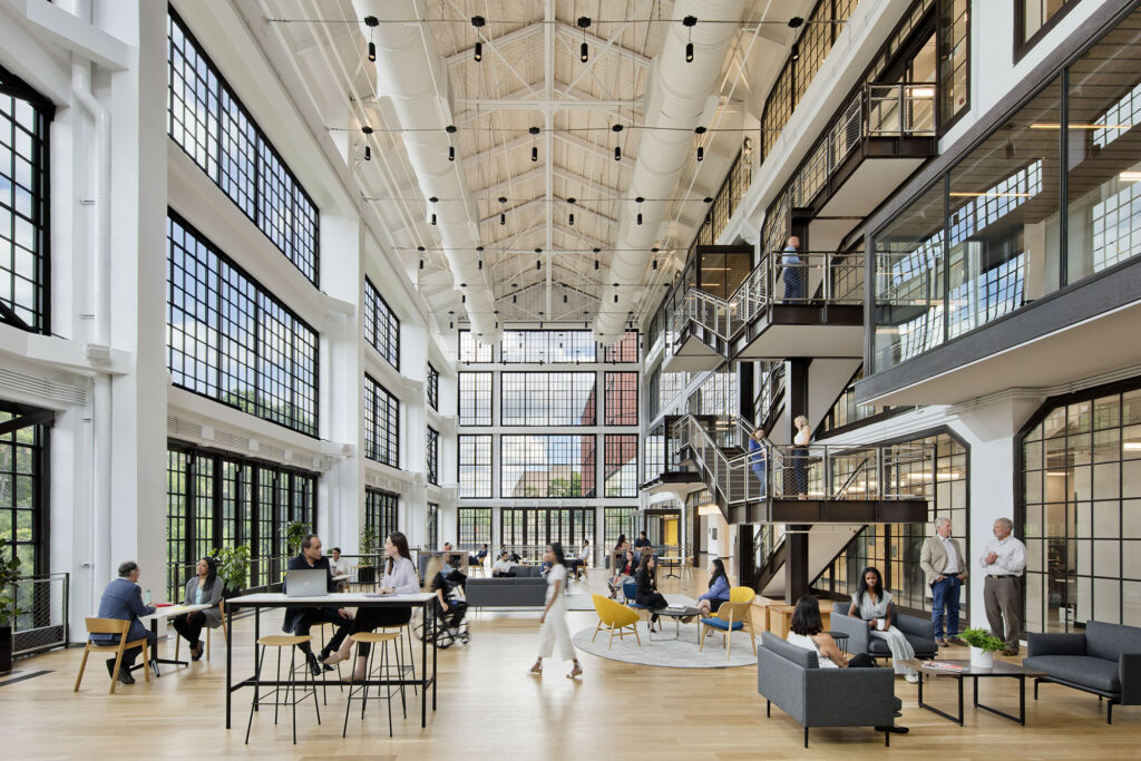 An open-concept modern office interior with high ceilings and large windows. Several people are engaged in conversations at tables, while others are seated in lounge areas. A staircase leads to an upper level.