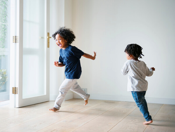 Two children running barefoot in a bright room with wooden flooring. One child has curly hair and wears a blue shirt and white pants. The other child wears a gray shirt and blue jeans.
