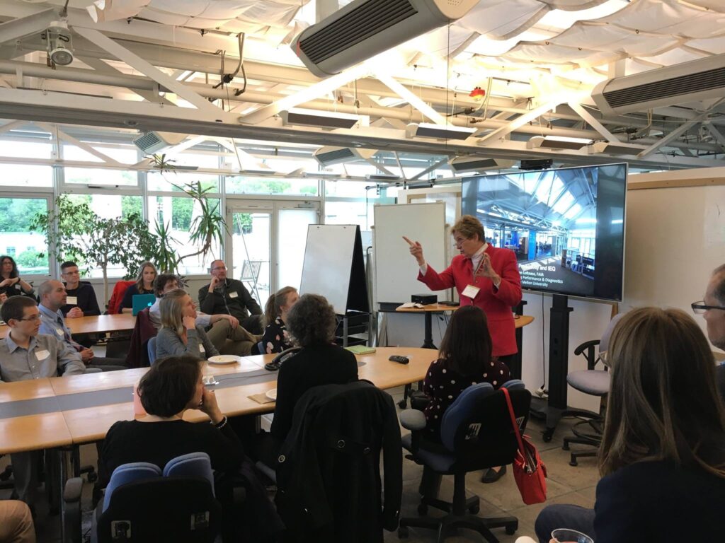 A woman in a red jacket speaks to an audience in a conference room. The room has large windows, plants, and a screen displaying an image. Attendees sit at a long table, listening attentively.