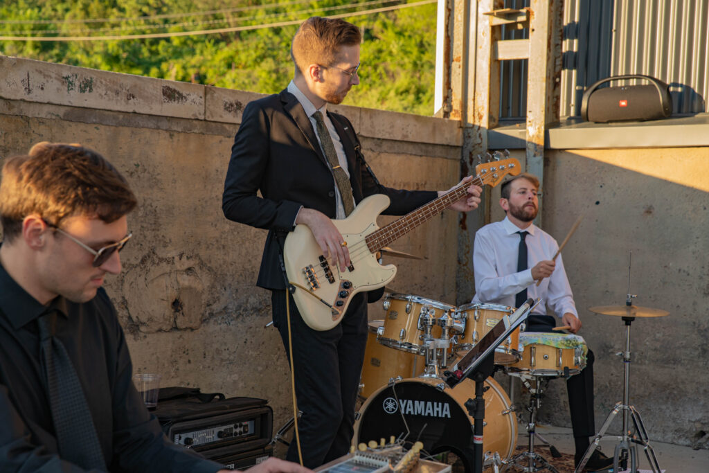 A three-member band performing outdoors. One musician plays a bass guitar, another is on drums, and the third plays a keyboard.