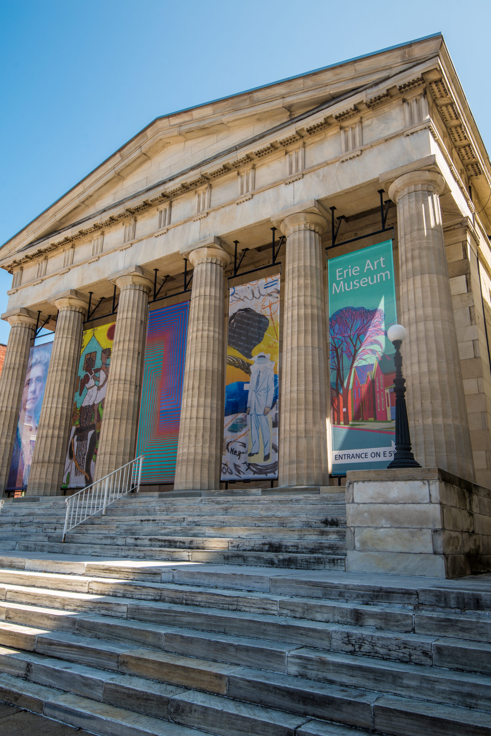 The exterior of the Erie Art Museum features a classical architectural style with tall columns and steps leading to the entrance. Colorful banners display various artworks on the front facade.