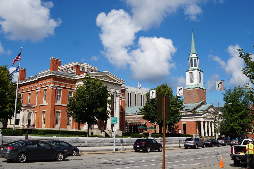 A view of a historic building with red brick exterior, greenery in front, and a clock tower with a green spire. Cars are parked along the street. The sky is partly cloudy.