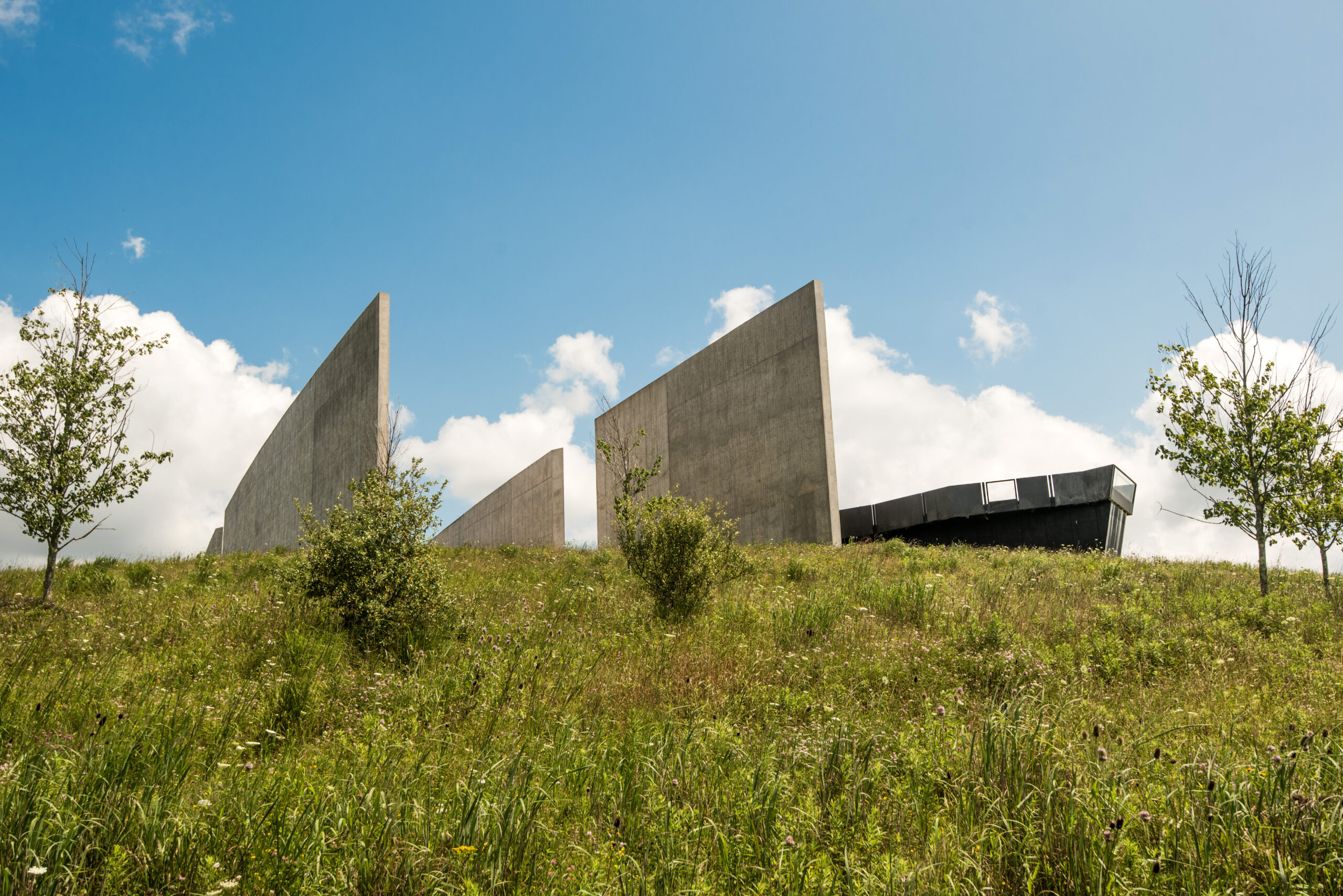 A grassy hill with a modern concrete memorial featuring angular panels, under a blue sky with clouds. Small trees are visible in the foreground.