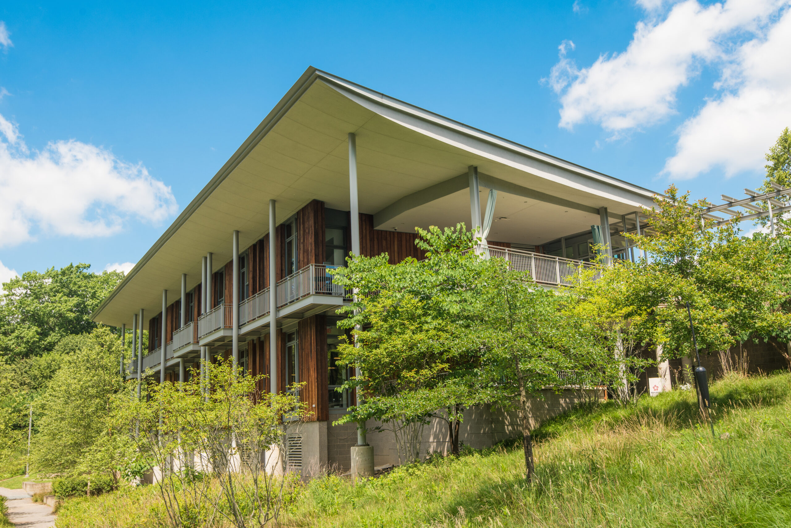 A modern two-story building with large windows and a sloped roof, surrounded by green trees and grass. The structure features wooden accents and a patio area on the upper level.