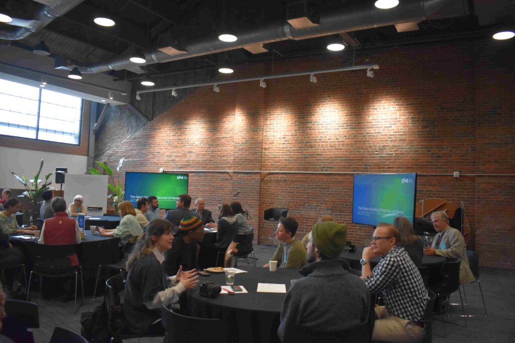 A group of people engaged in discussion at tables in a modern meeting space. Two screens are displaying information in front of a brick wall. Potted plants are visible, and participants are seated in black chairs.