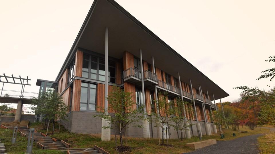 Modern building with a wide roof, featuring wooden panels and large windows. Surrounding area includes trees and pathways leading to the entrance.