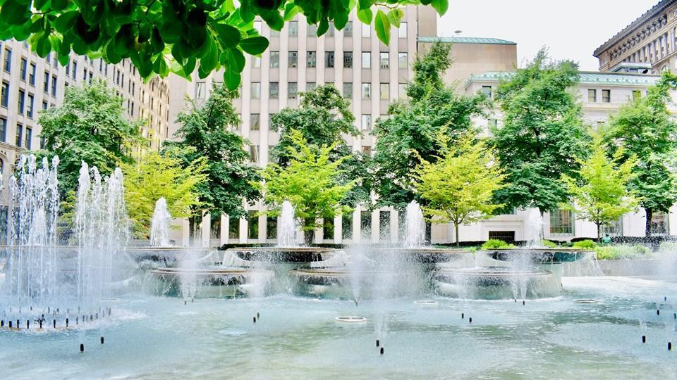 A public plaza featuring multiple water fountains surrounded by green trees and tall buildings. Mist rises from the fountains, creating a refreshing atmosphere.