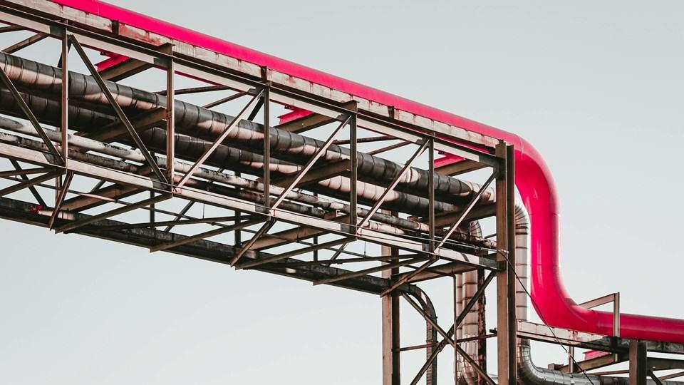 A red industrial pipe twisting along a metal framework against a pale sky. The structure shows a combination of metal beams and additional piping, emphasizing an industrial design.