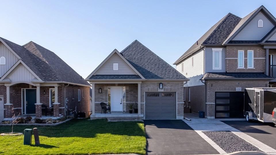 A residential neighborhood featuring three houses. The center house has a gray stone facade and a garage. The left house has a brick exterior, and the right house is larger with a light-colored facade. A lawn is present in front of the houses.