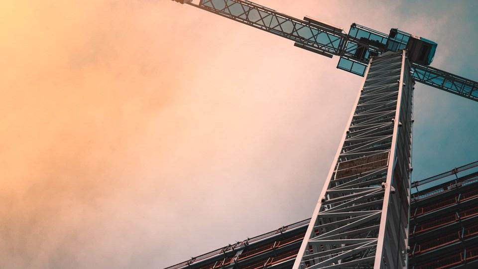 A construction crane reaching upward against a cloudy sky. The crane's platform and structural elements are visible, with a gradient of orange and blue in the background.