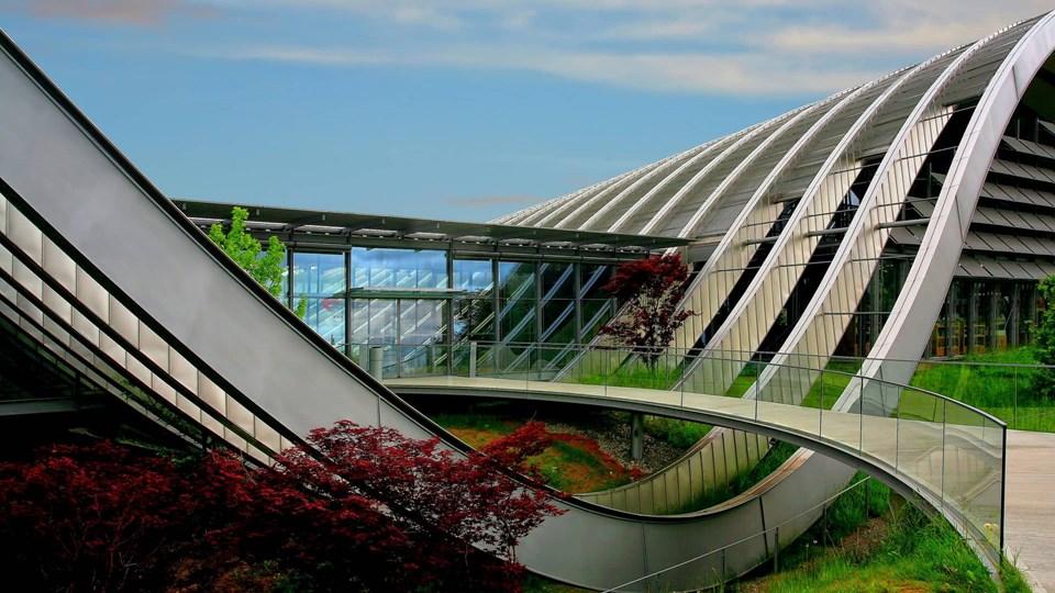 A modern building with curved architectural features, glass panels, and landscaped greenery. The structure has a pathway that winds between vegetation, highlighting its contemporary design.