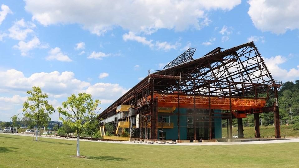 A partially constructed building with an exposed framework and bright orange elements. Surrounding green grass and a few small trees are visible. The sky is partly cloudy.