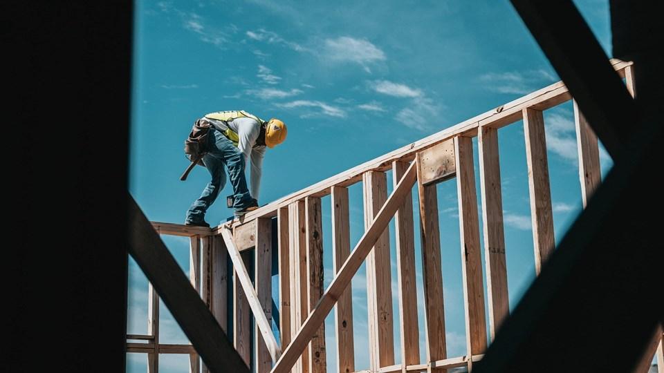 A construction worker wearing a yellow hard hat and safety gear stands on a wooden frame of a building under a blue sky. The view is framed by dark structural beams.