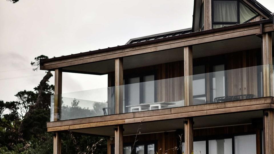 A modern two-story building with wooden siding and large glass windows, featuring a balcony with a glass railing. The roof has a slight slope and a small gable. Trees are visible in the background.