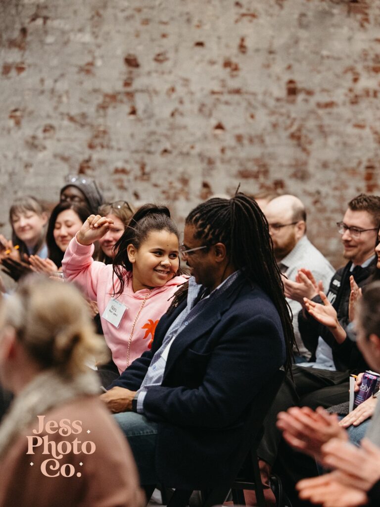 A girl in a pink sweatshirt is smiling and raising her hand while sitting next to a man in a dark jacket at an event. Audience members are clapping in the background, with a brick wall behind them.