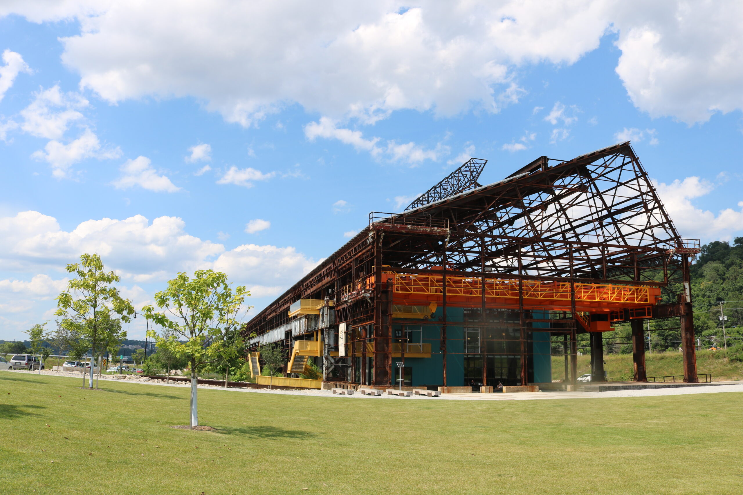 An industrial building with a steel frame and colorful orange and yellow exterior, set in a grassy area under a blue sky with clouds. Nearby trees and a sidewalk are visible.