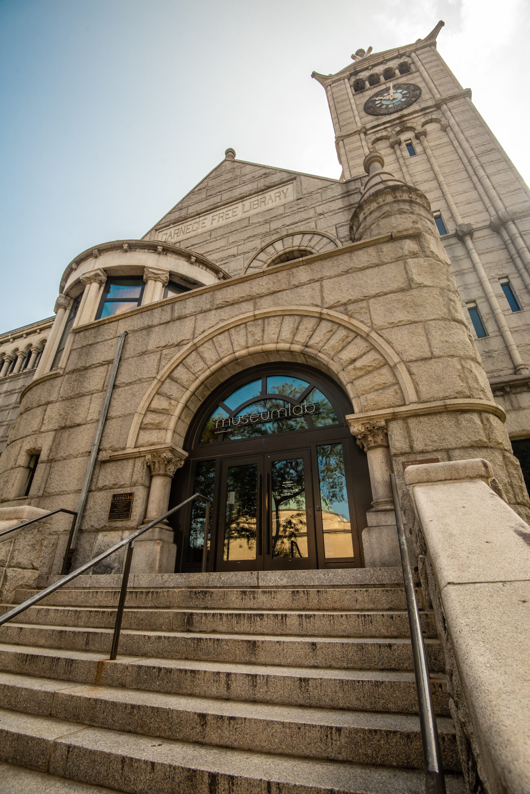 MuseumLab Entrance. The entrance of a stone building with a large clock tower, featuring arched doorways and staircases leading to the entry door with visible glass panels.