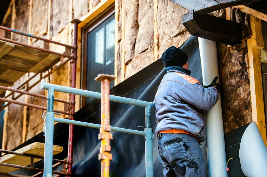 A construction worker installing a pipe on the exterior of a building. He is on a scaffold, wearing a dark hat and a gray jacket, with insulation material visible on the walls.