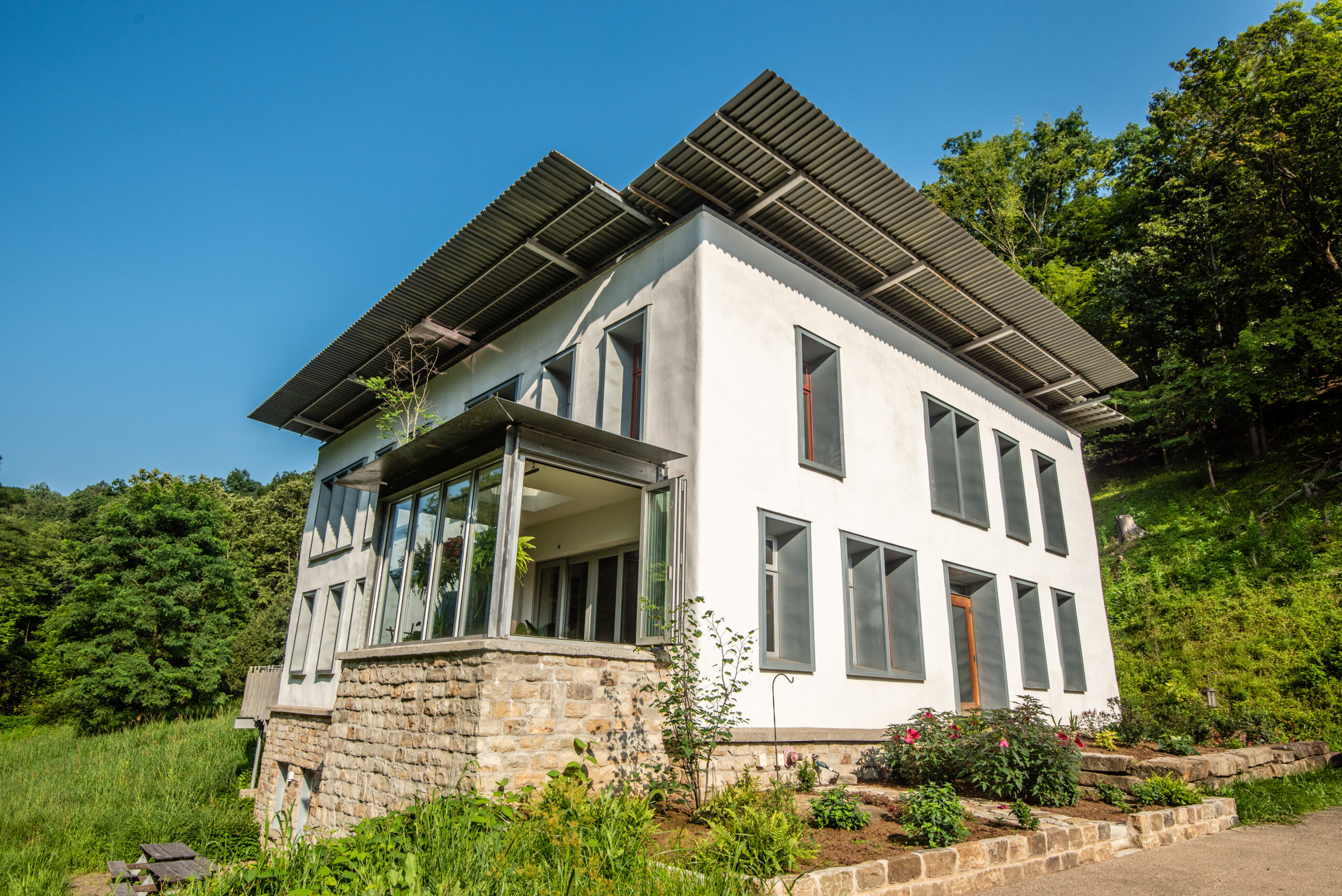 A modern two-story building with large windows and a stone base, surrounded by greenery. The structure features a sloping roof with solar panels and a glass-enclosed porch area.