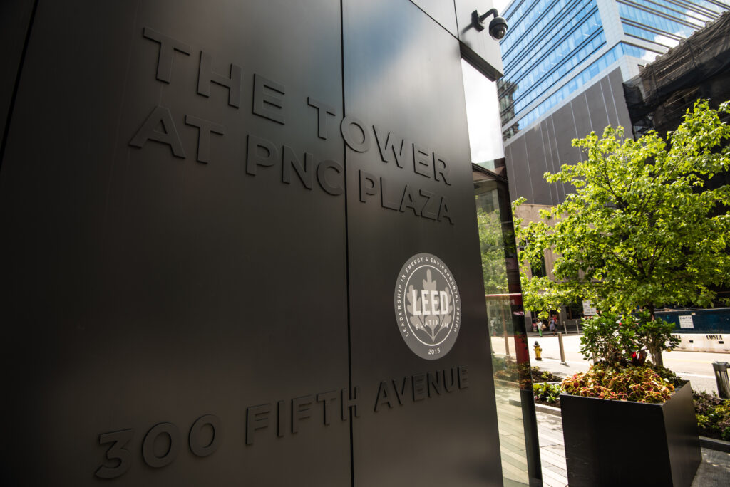 A close-up of a building sign that reads "THE TOWER AT PNC PLAZA" and "300 FIFTH AVENUE". There is a circular LEED certification logo visible. Surrounding area includes greenery and part of the building's facade.