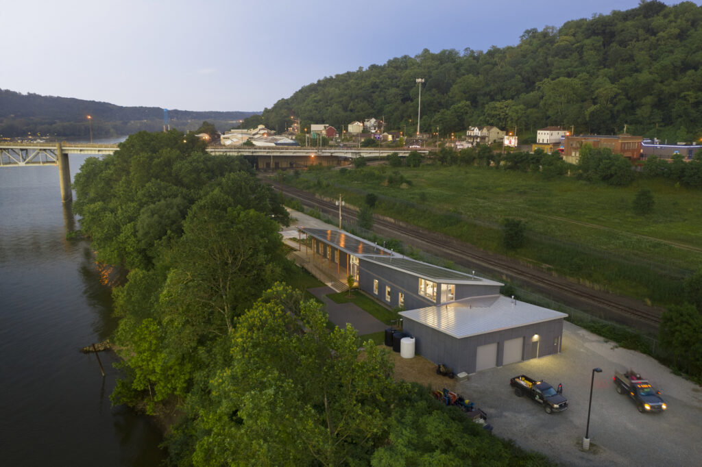 An aerial view of a modern building near a river, surrounded by trees and grassy areas. There are parked cars near the building and a bridge in the background.