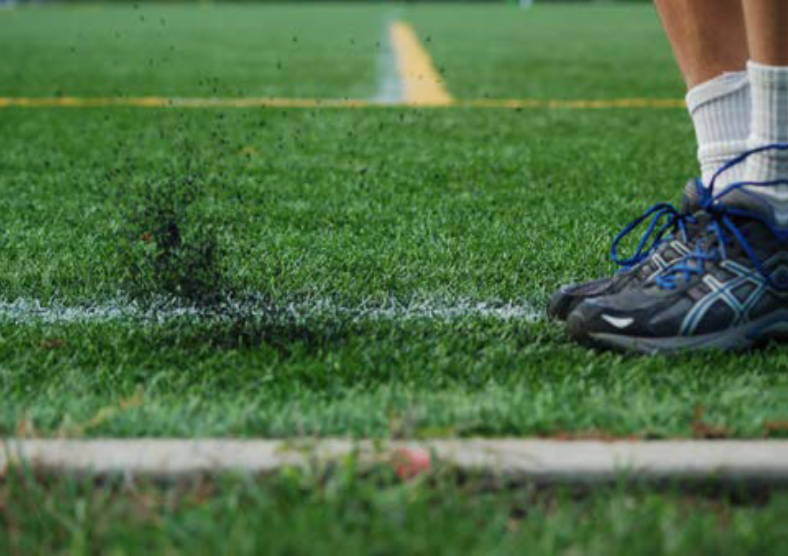 A close-up of a person's athletic shoes on a grass field, with dust or rubber particles dispersing as the foot strikes the ground near a white line.